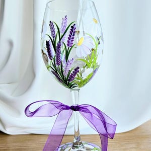 Könnte beinhalten: Ein klares Weinglas, handbemalt mit Lavendel und weißen Gänseblümchen. Ein lila Band ist um den Stiel gebunden. Der Fuß des Glases ist ebenfalls mit weißen Blumen verziert. Das Glas ist etwa 20 cm hoch.
