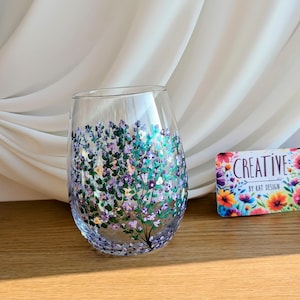 Könnte beinhalten: Ein klares Weinglas aus Glas mit handbemaltem Design aus lila und grünen Blumen. Das Glas steht auf einer Holzoberfläche. Der Text "CREATIVE BY KAT DESIGN" ist auf einer Visitenkarte im Hintergrund sichtbar.