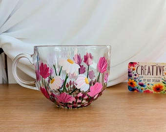 Könnte beinhalten: Eine klare Glas-Tasse, handbemalt mit rosa und weißen Blumen. Die Tasse hat eine abgerundete Form und einen Henkel. Die Blumen sind realistisch gemalt und beinhalten Gänseblümchen und Tulpen.