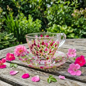 May include: A clear glass teacup and matching saucer decorated with hand-painted pink and red floral patterns. The set features delicate stems and leaves in green, with small blossoms scattered across the surface of both the cup and the saucer.