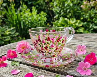 Könnte beinhalten: Eine Teetasse mit passender Untertasse aus klarem Glas, verziert mit handgemalten rosa und roten Blumenmustern. Das Set zeigt zarte grüne Stängel und Blätter, wobei kleine Blüten über die Oberfläche der Tasse und der Untertasse verteilt sind.