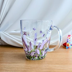 May include: A clear glass mug with a handle, featuring a hand-painted design of lavender flowers and butterflies in shades of purple, pink, and green.