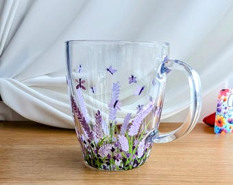 May include: A clear glass mug with a handle, featuring a hand-painted design of lavender flowers and butterflies in shades of purple, pink, and green.