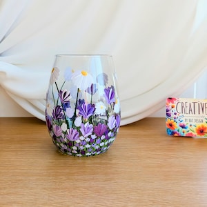 Könnte beinhalten: Handbemaltes Weinglas mit einem floralen Design mit violetten und weißen Blumen. Das Glas ist klar und hat eine runde Form.