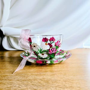 Op de afbeelding: Een heldere glazen theekop en schotel set versierd met handgeschilderde bloemen in wit, roze en rood. Een roze lint is aan het handvat gebonden. De set staat op een houten oppervlak.