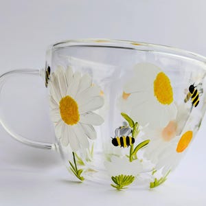 Op de afbeelding: Een heldere glazen mok met een ontwerp van witte madeliefjes en bijen. De mok heeft een handvat en is versierd met gele en groene accenten.