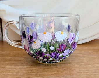 Könnte beinhalten: Eine transparente Glas-Tasse mit einem bemalten Blumendesign. Die Blumen sind weiß, lila und lavendelfarben mit gelben Zentren. Die Tasse hat einen Henkel und ist für Heißgetränke geeignet.