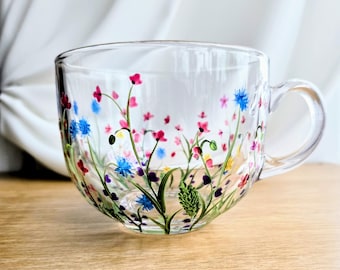 Könnte beinhalten: Eine transparente Teetasse mit Henkel, handbemalt mit einem farbenfrohen Blumenmuster. Die Blumen sind rosa, blau und lila, mit grünen Stielen und Blättern. Die Tasse steht auf einer hellbraunen Holzoberfläche.
