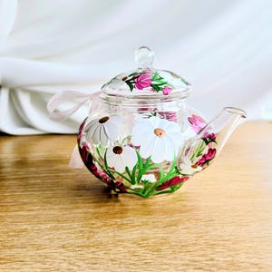 Può includere: Una teiera in vetro trasparente con coperchio, ornata da margherite bianche dipinte a mano, fiori rosa e foglie verdi. Un nastro rosa è legato attorno alla teiera. La teiera è su una superficie di legno.