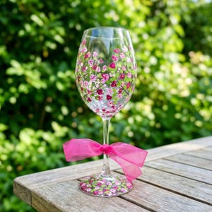 Puede incluir: Una copa de vino de cristal transparente con flores pintadas a mano en tonos rosa y morado con hojas verdes en el cáliz y la base. Un lazo de cinta rosa brillante y translúcida está atado alrededor del tallo de la copa sobre una superficie de madera.