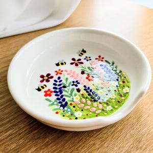 May include: A small, white ceramic dish with a hand-painted floral design. The design features colourful flowers, green grass, and two yellow and black bees. The dish has a small, curved handle and is set on a wooden surface.