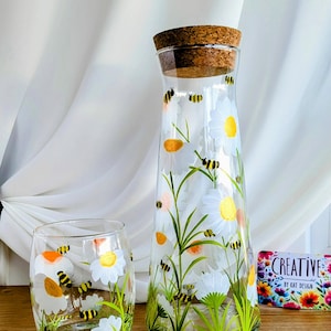 Op de afbeelding: Een set van twee glazen containers met een handgeschilderd ontwerp van witte madeliefjes en gele bijen. De containers hebben een kurken stop en zijn perfect om dranken te serveren.