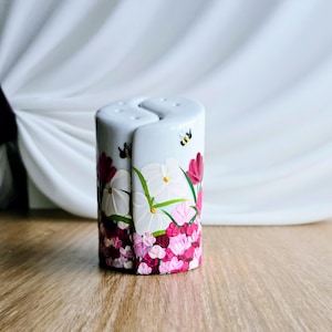 Puede incluir: Un juego de salero y pimentero de cerámica blanca. El juego es cilíndrico con un diseño entrelazado único. Está pintado a mano con un patrón floral en tonos de rosa, blanco y verde, con un pequeño acento de abeja. El juego mide aproximadamente 10 cm de alto.