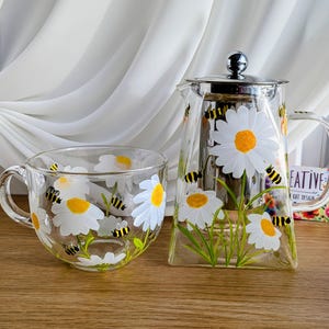 Può includere: Una teiera di vetro e una tazza abbinata con un design dipinto a mano di margherite bianche e centri gialli con api nere e gialle. La teiera ha un coperchio e un filtro in acciaio inossidabile.