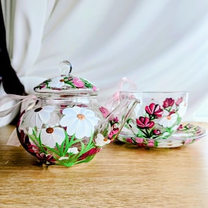 Puede incluir: Un juego de tetera y taza de té de vidrio transparente decorado con margaritas blancas y flores rosas pintadas a mano. La tetera tiene tapa y asa, y la taza se apoya en un platillo a juego. El juego está sobre una superficie de madera.
