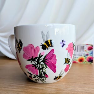 Könnte beinhalten: Weiße Keramiktasse mit handbemaltem Design aus rosa Blumen und Hummeln. Die Tasse hat eine abgerundete Form und einen Henkel.