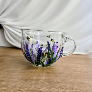 Può includere: Una tazza da tè in vetro trasparente con manico, dipinta a mano con fiori di lavanda nelle tonalità del viola e del verde. Piccole api gialle e nere sono sparse intorno alla tazza, creando un design stravagante. La tazza è su una superficie di legno.