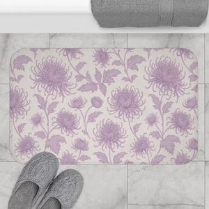 Könnte beinhalten: Ein rechteckiger Badteppich mit einem floralen Muster aus hellvioletten Chrysanthemenblüten und Blättern auf cremefarbenem Hintergrund. Der Teppich liegt auf einem gefliesten Boden, mit einem Paar grauer Hausschuhe unten.
