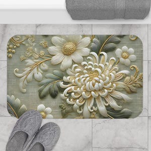 May include: A beige bath mat with a floral design featuring white and gold flowers. The flowers are stylized and have a 3D effect.
