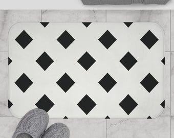May include: A rectangular white bath mat with a repeating pattern of black diamond shapes. Gray slippers are at the bottom of the mat. A gray towel is at the top of the image.
