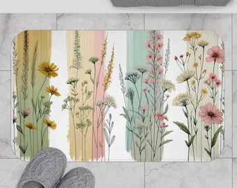 Könnte beinhalten: Eine weiße Badematte mit einem farbenfrohen Blumendesign. Das Design zeigt eine Vielzahl von Blumen und Pflanzen in Rosa-, Gelb- und Grüntönen. Die Blumen sind in einem vertikalen Muster angeordnet, wobei die Stiele vom Boden der Matte nach oben reichen. Der Hintergrund der Matte ist weiß mit einem dezenten Pinselstrichmuster.