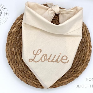 May include: A cream-colored dog bandana with the name "Louie" embroidered in beige thread. The bandana is knotted at the top and rests on a woven, circular mat. The text "FONT 6" and "BEIGE THREAD" are visible.
