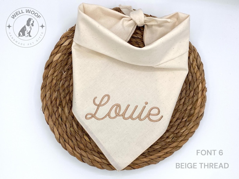 May include: A cream-colored dog bandana with the name "Louie" embroidered in beige thread. The bandana is knotted at the top and rests on a woven, circular mat. The text "FONT 6" and "BEIGE THREAD" are visible.