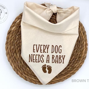 May include: A white cotton bandana with brown text that reads "Every Dog Needs A Baby" and two brown footprints. The bandana is tied in a knot at the top and is lying on a woven brown surface.