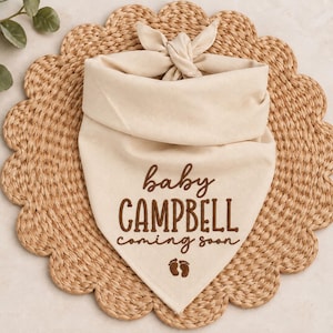 May include: A cream-colored triangular pet bandana featuring a tie-top closure. The fabric is embroidered with dark brown text that reads "baby CAMPBELL coming soon" above a small graphic of two baby footprints centered at the bottom point.