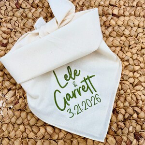 May include: Cream-colored dog bandana with the names "Lele & Garrett" and the date "3.21.2026" embroidered in green. The bandana is tied at the top and rests on a woven, light brown surface.