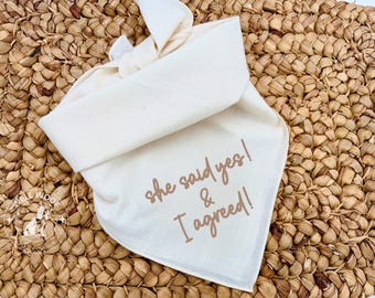 Peut inclure: Bandana crème pour chien avec l'inscription "she said yes! & I agreed!" en script marron. Le bandana est plié et noué, posé sur une surface tissée brun clair. Accessoire de mariage.