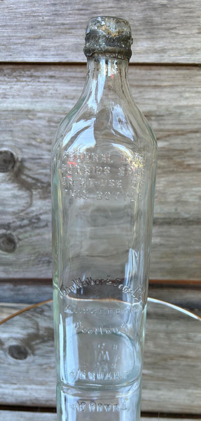 May include: Clear glass bottle with a metal cap. The bottle has embossed text that reads "FEDERAL LAW FORBIDS SALE OR RE-USE OF THIS BOTTLE" and "John Walker & Sons". The bottle is 1/5 quart.
