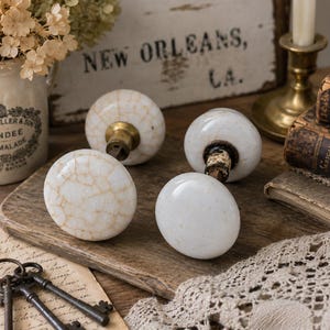 May include: Four antique white ceramic door knobs with brass hardware, arranged on a wooden cutting board. Two knobs have a cracked, aged appearance. The background includes vintage books, a candle holder, and a sign that says "New Orleans, LA."
