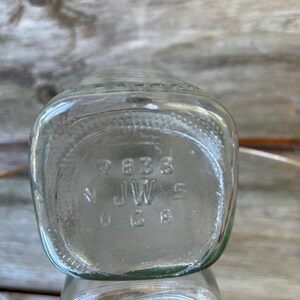 May include: Clear glass bottle with a square base. The bottom of the bottle has embossed text including the numbers "838", "JW", and "5". The bottle is partially visible, with a blurred wooden background.