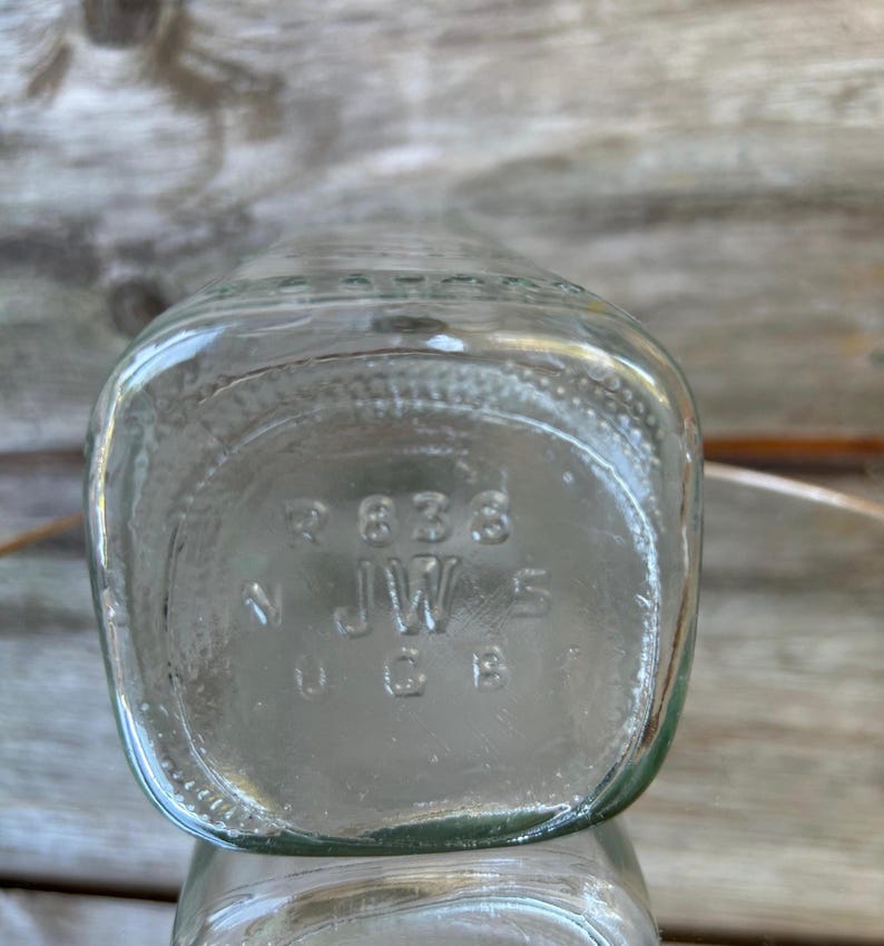 May include: Clear glass bottle with a square base. The bottom of the bottle has embossed text including the numbers "838", "JW", and "5". The bottle is partially visible, with a blurred wooden background.