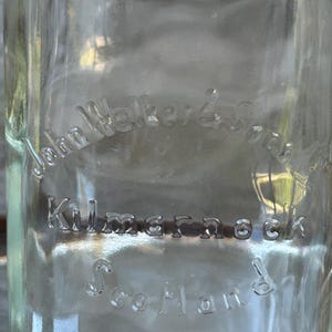 May include: Clear glass bottle with embossed text. The text reads "John Walker & Sons, Kilmarnock, Scotland". The bottle is empty and appears to be vintage. The glass has a slight green tint.