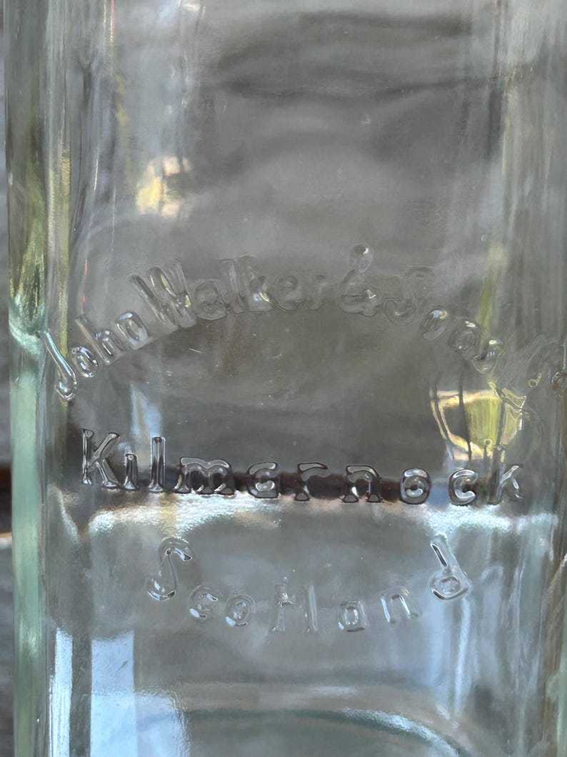 May include: Clear glass bottle with embossed text. The text reads "John Walker & Sons, Kilmarnock, Scotland". The bottle is empty and appears to be vintage. The glass has a slight green tint.