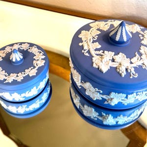May include: Two blue ceramic trinket boxes with white relief grape vine designs. The boxes have a round shape with lids and a decorative finial on top. The boxes are displayed on a reflective surface.