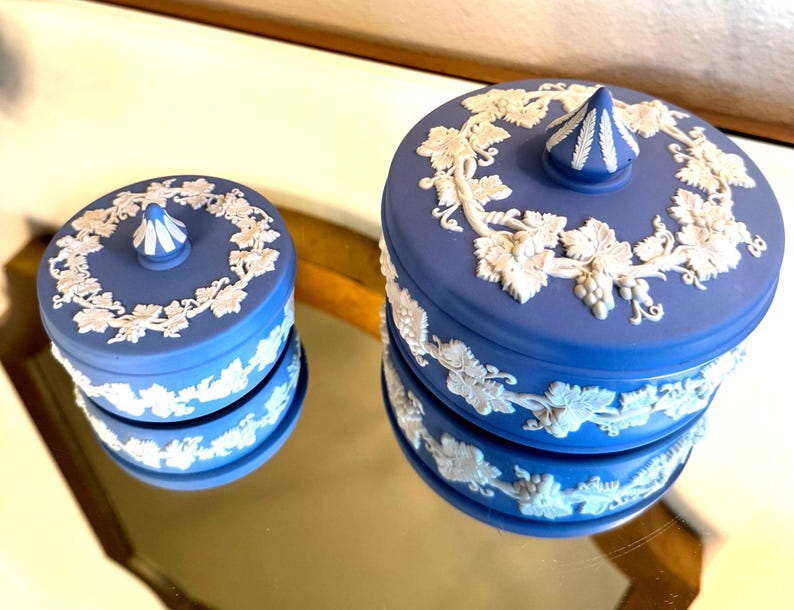 May include: Two blue ceramic trinket boxes with white relief grape vine designs. The boxes have a round shape with lids and a decorative finial on top. The boxes are displayed on a reflective surface.