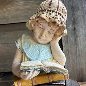 May include: A vintage-style statue of a child reading a book. The statue features a girl wearing a hat and a blue collar, with her head resting on her hand. The book is open, and the statue is placed on top of a stack of books.