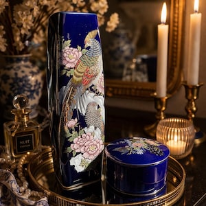 May include: A tall, cobalt blue vase and a small round box with matching peacock and floral designs. The vase and box are on a gold-toned tray. The vase features a detailed illustration of a colorful bird and flowers.