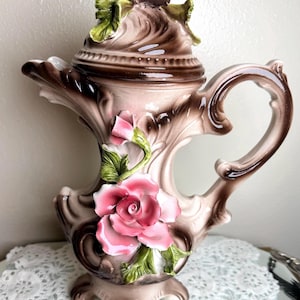 May include: A vintage ceramic teapot with a lid, featuring a pink and brown floral design. The teapot has a large pink rose and green leaves, with a decorative handle and base. The teapot is a decorative item.
