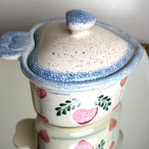 May include: A ceramic heart-shaped casserole dish with a lid. The dish is white with blue trim and a speckled pattern. Pink hearts and green leaves decorate the exterior. The lid has a blue knob.