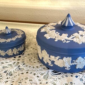 May include: Two blue ceramic lidded jars with white relief grape vine designs. The jars are different sizes, with a smaller one on the left and a larger one on the right. The lids have a conical finial.
