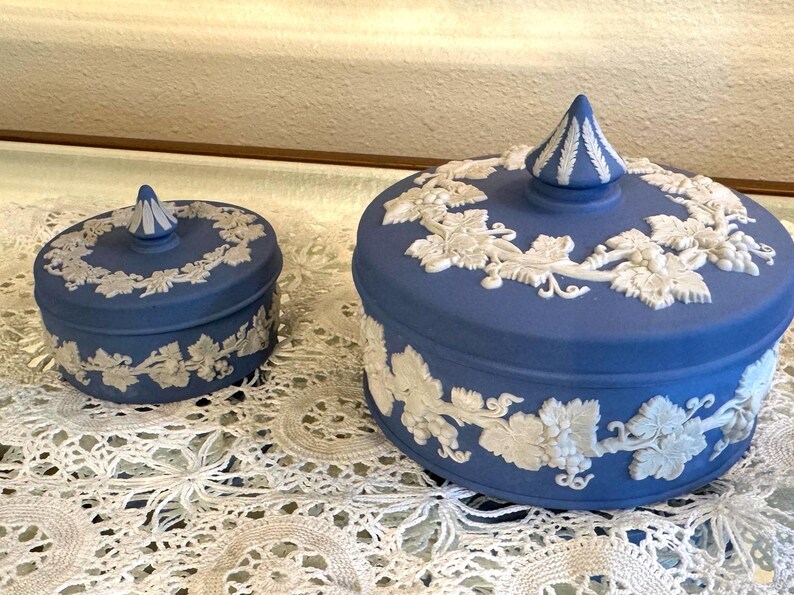 May include: Two blue ceramic lidded jars with white relief grape vine designs. The jars are different sizes, with a smaller one on the left and a larger one on the right. The lids have a conical finial.