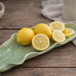 May include: A green leaf-shaped ceramic serving platter holds several fresh lemons, some sliced. The lemons are bright yellow, contrasting with the platter's pale green. A bowl of green olives sits nearby on a wooden surface.