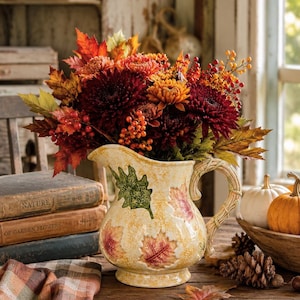 May include: A cream-colored ceramic pitcher with a textured surface featuring embossed autumn leaf motifs in green, red, and orange. The pitcher has a braided handle and is filled with a vibrant bouquet of dark red chrysanthemums, orange berries, and maple leaves.