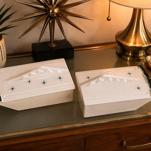 May include: Two rectangular white ceramic boxes with lids, featuring a raised geometric roof-like design and black starburst patterns on the top. Each box has dark green accents on the side edges, creating a cohesive set of storage containers.