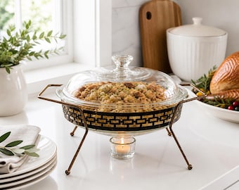 Pode incluir: Um aquecedor de alimentos de vidro e metal com tampa de vidro transparente, cheio de comida cozida. O aquecedor tem uma base com padrão preto e dourado e é suportado por pernas de cor bronze. Uma vela acesa está colocada por baixo.