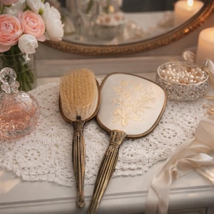 May include: A vintage vanity scene featuring a brush, hand mirror, and perfume bottle. The brush and mirror have ornate gold handles. A vase of pink and white flowers, a candle, and a bowl of pearls complete the elegant display.